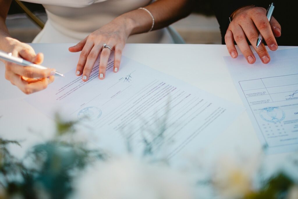 Close-up van handen met pennen die de huwelijksakte ondertekenen op een witte tafel, met bloemen op de voorgrond